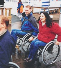 A man and a woman who both use wheelchairs wheeling along a boardwalk.